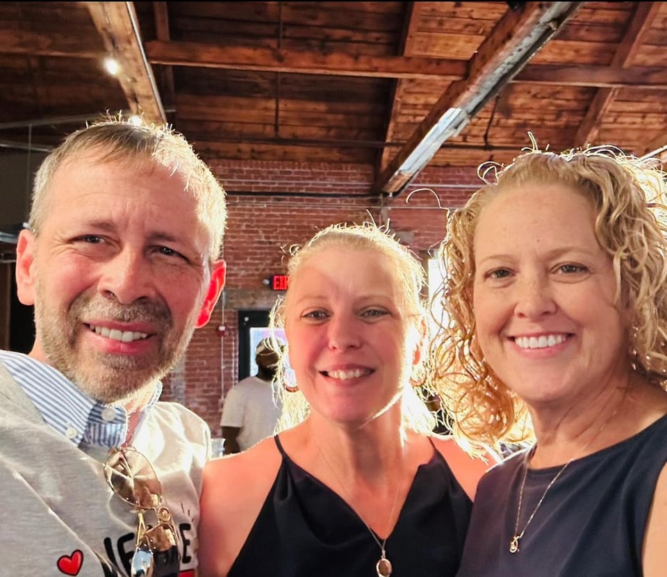 A man and two women smile for a close-up selfie beneath a wooden ceiling.