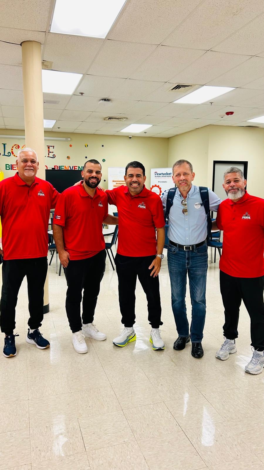 A group of men poses together in a brightly lit hallway, with two wearing red shirts.