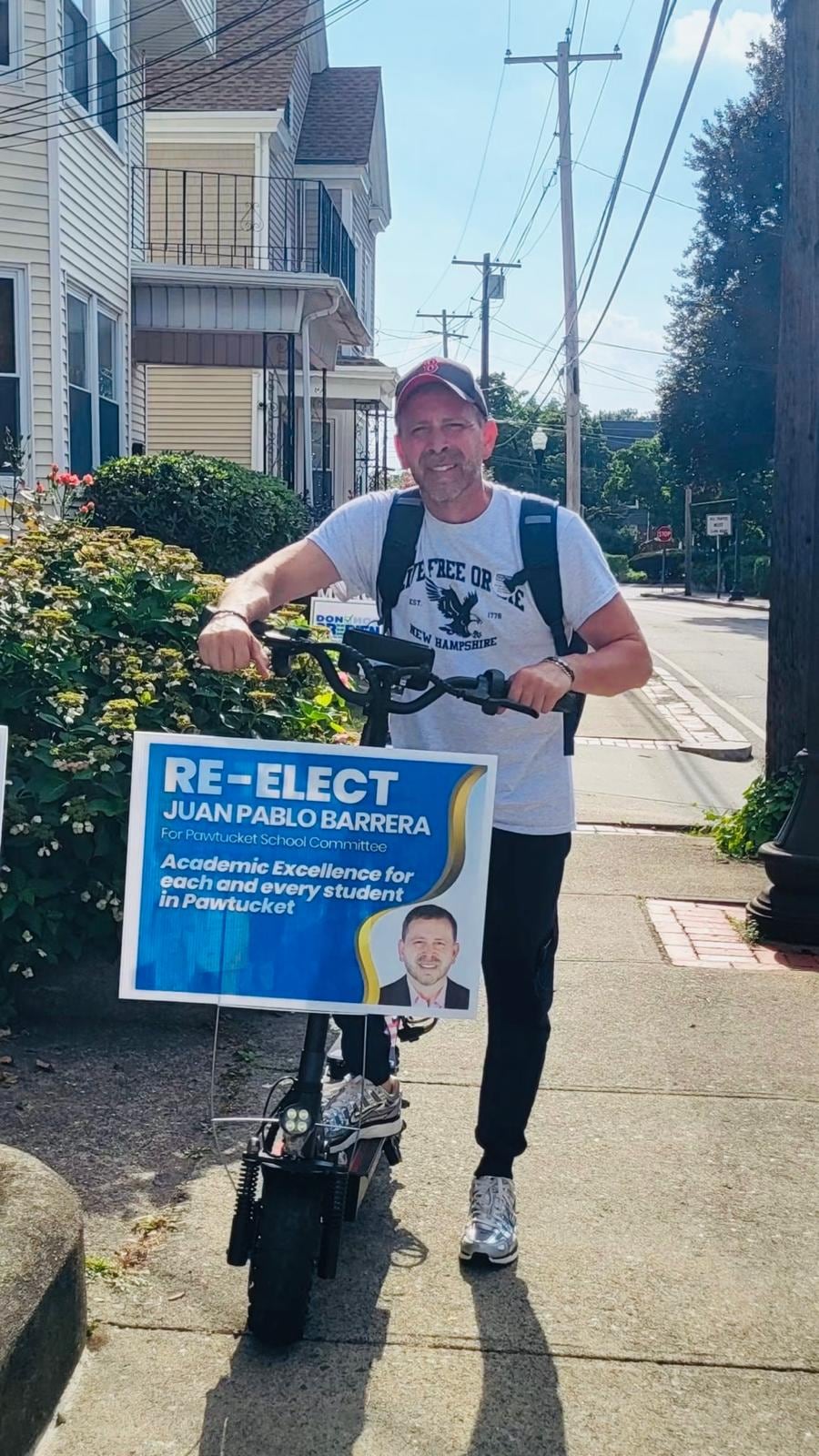 A man stands on a sunny sidewalk holding a blue “Re-elect Juan Pablo Barrera” sign.
