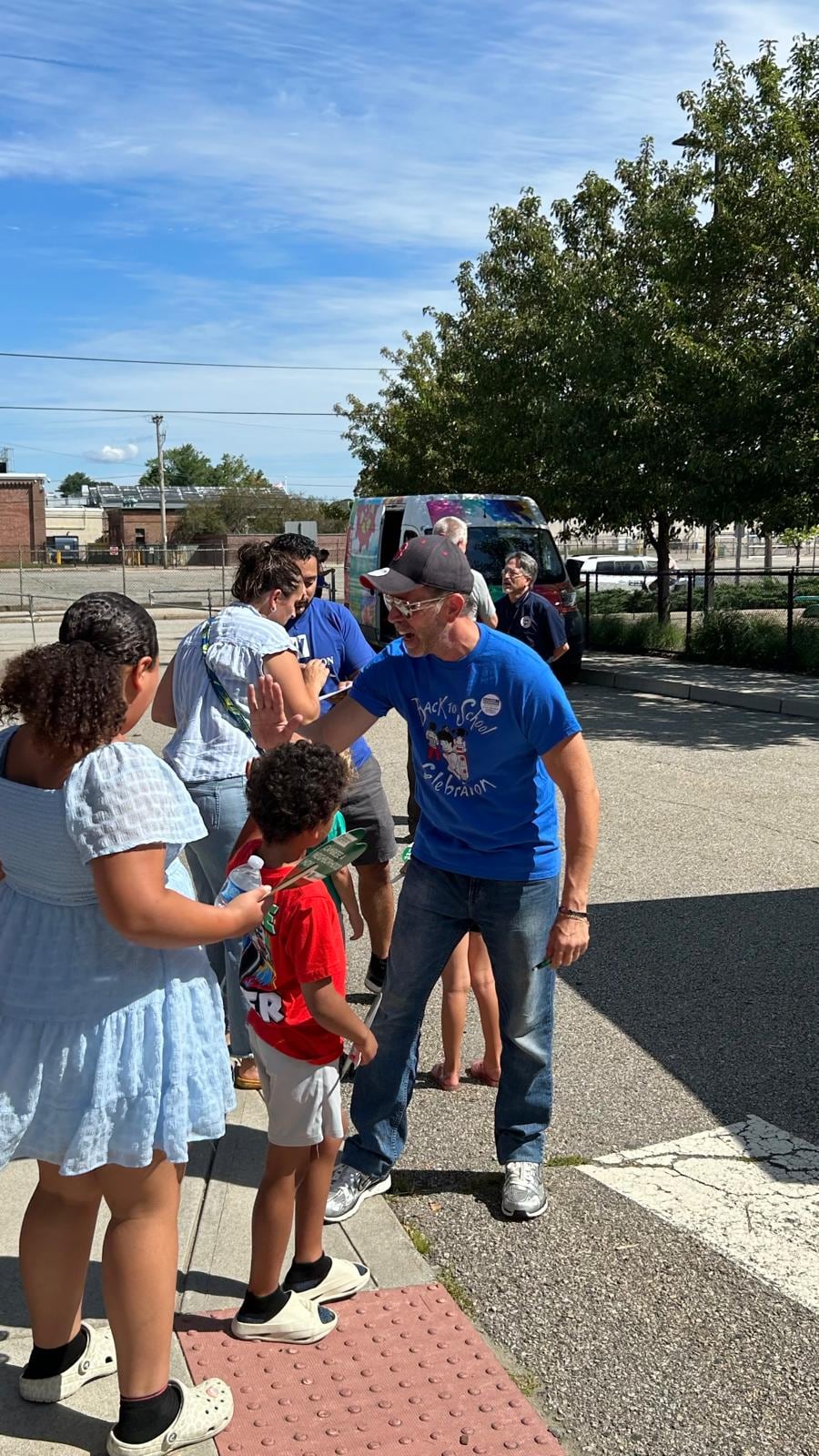 A man in a blue shirt bends toward a child while several people gather around them outdoors.