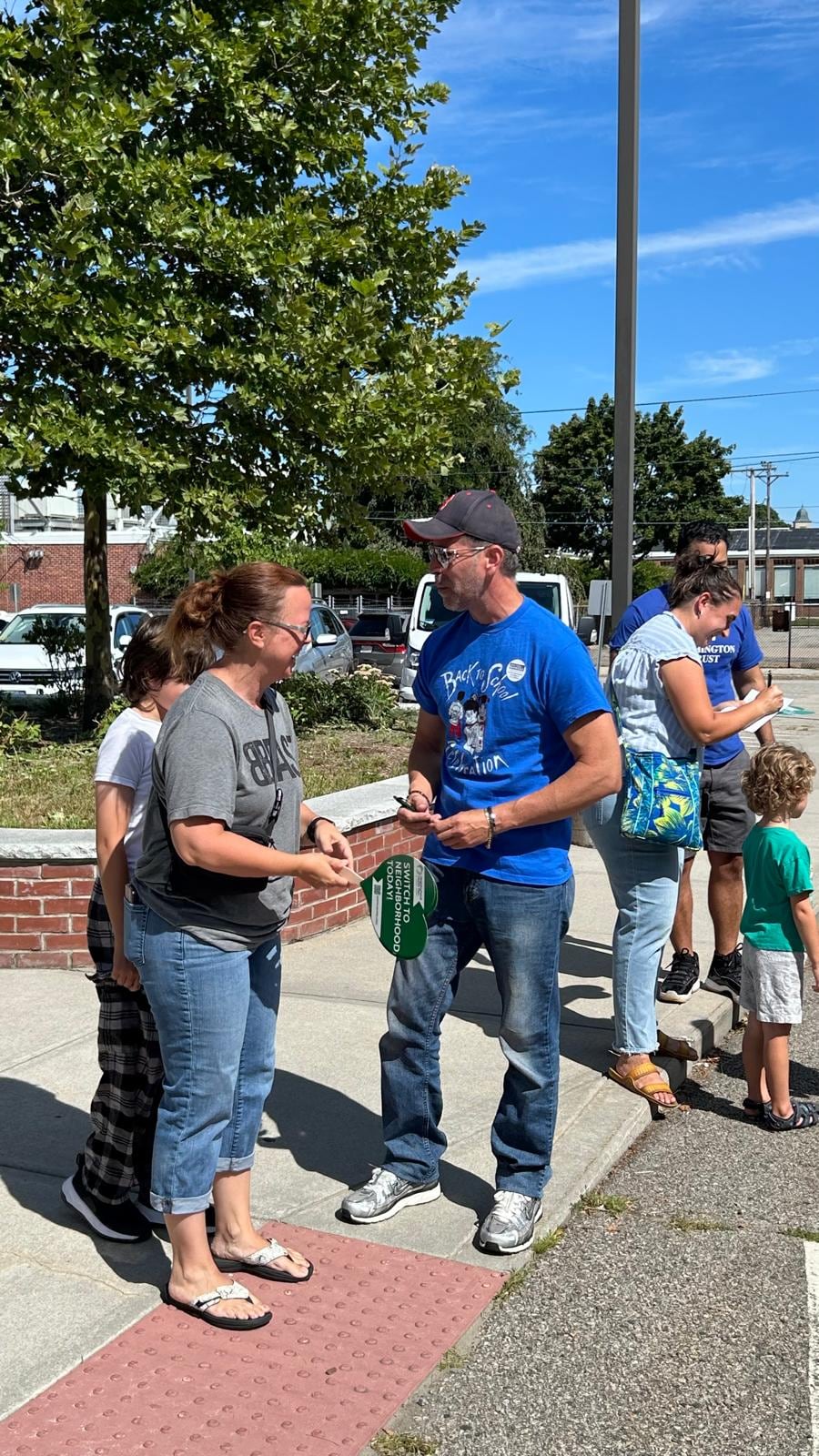 A man in a blue shirt speaks with a woman as other people stand nearby in a sunny parking lot.
