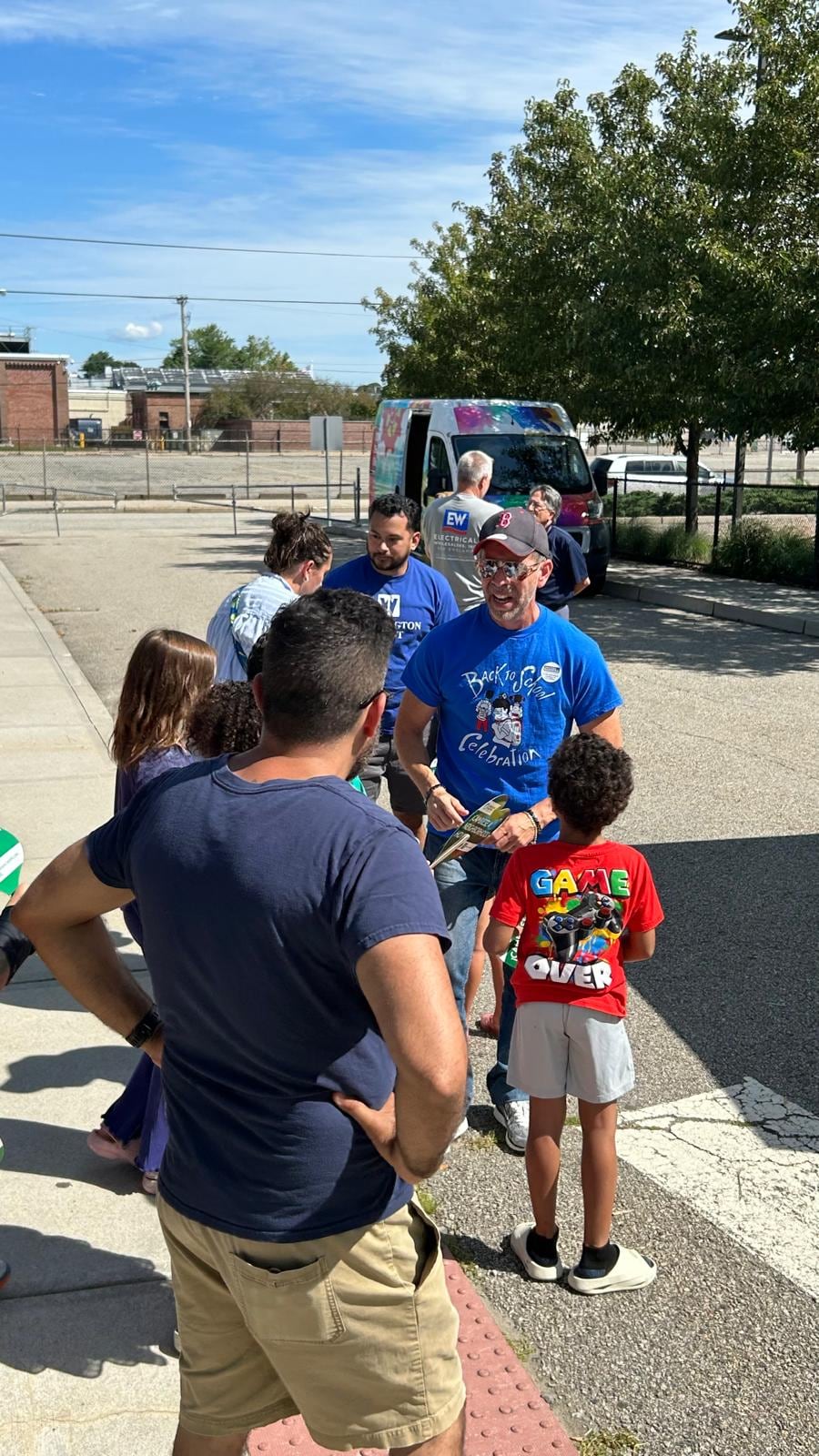 Adults and children gather around a man in a blue shirt in a sunny parking lot.