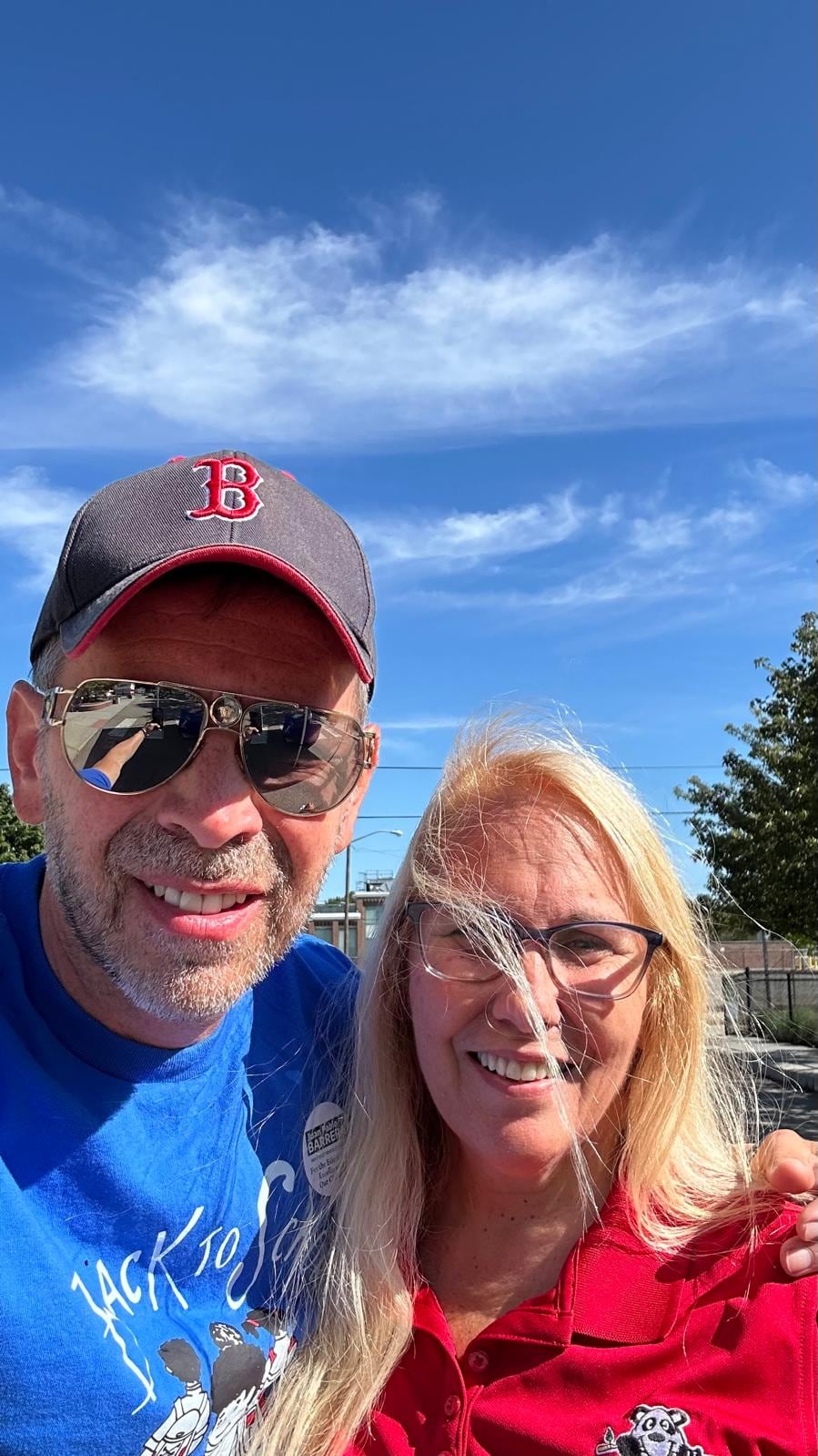 A man in a red cap and sunglasses smiles beside a woman wearing glasses in an outdoor selfie.