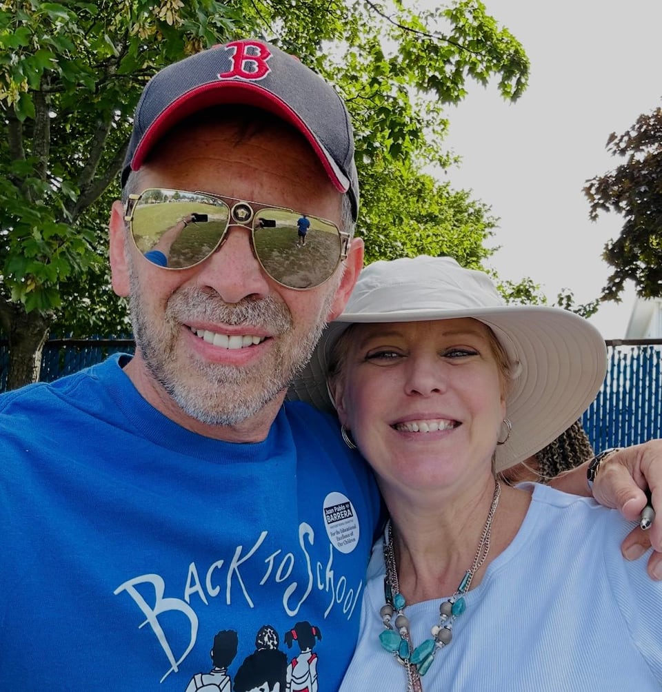 A man in a red cap and mirrored sunglasses smiles beside a woman in a wide-brimmed hat.