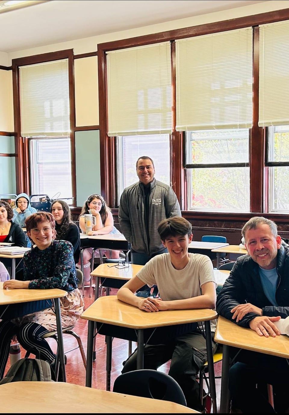 Several adults sit at classroom desks while two men stand near the back of the room.