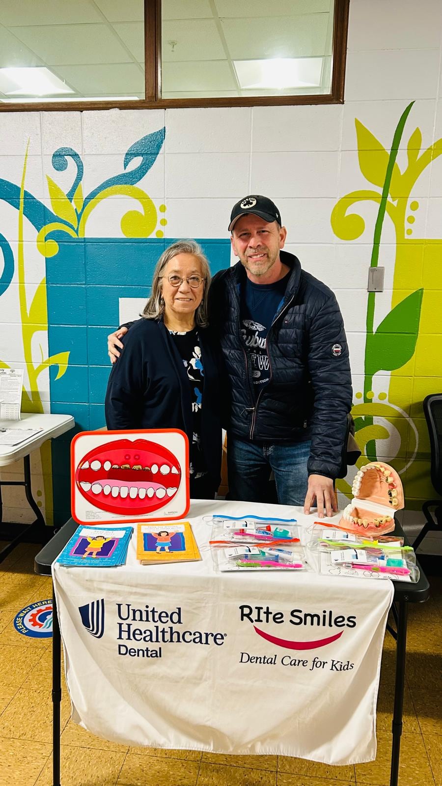 A woman and a man stand behind a table displaying signs, handouts, and small giveaway items.