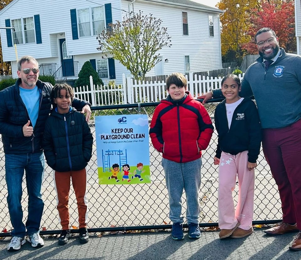 Six people pose beside a chain-link fence with a colorful “Keep Our City Beautiful” sign.