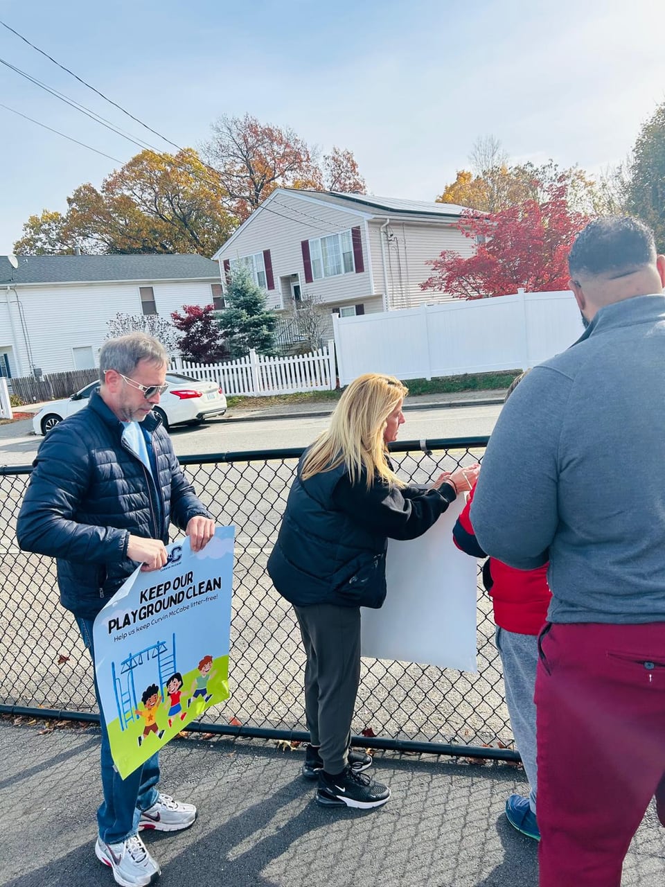 Three adults stand by a chain-link fence, with one holding a colorful printed sign.