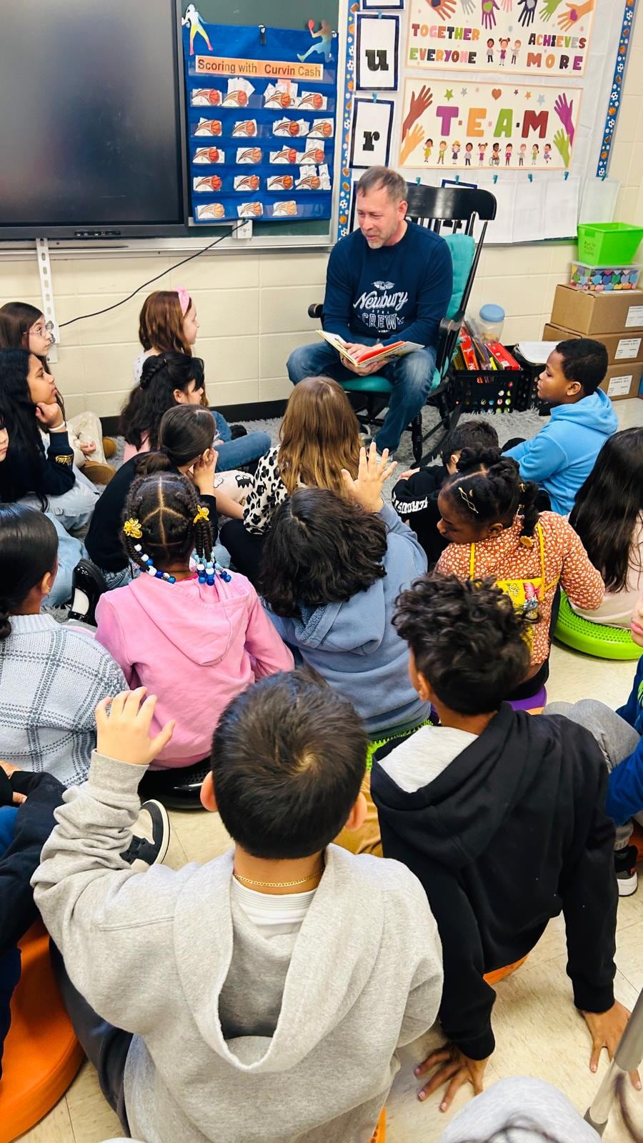 A man seated at the front of a classroom speaks to children sitting closely together on the floor.