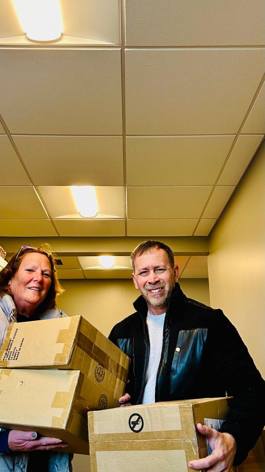 A woman and a man smile while holding two stacked cardboard boxes in a hallway.