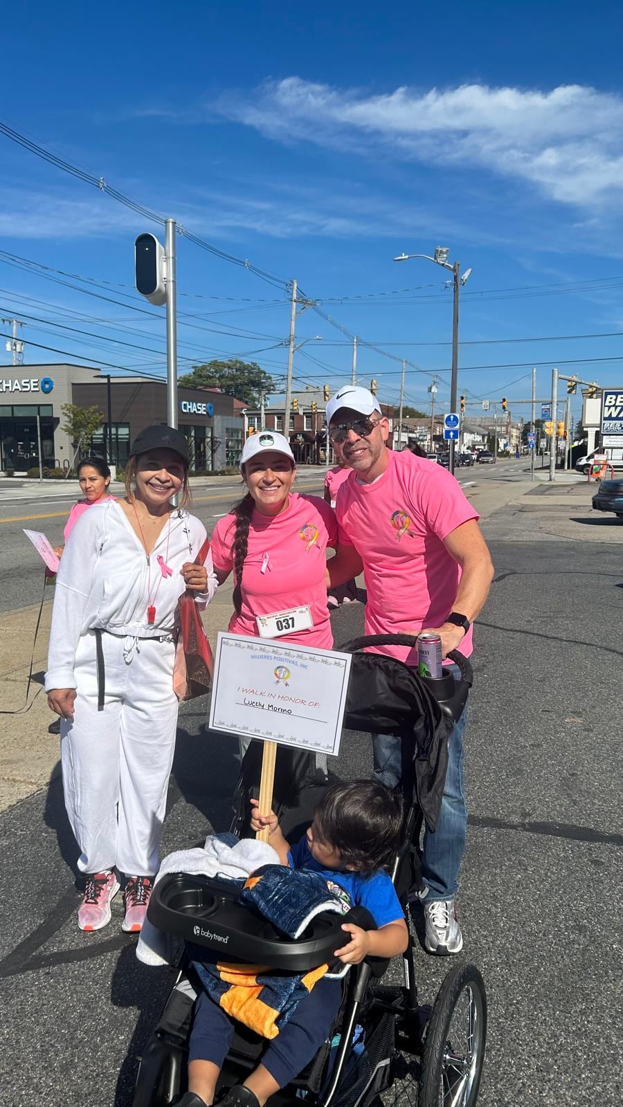 A group in pink and white clothing poses outdoors with a printed sign and bags at their feet.