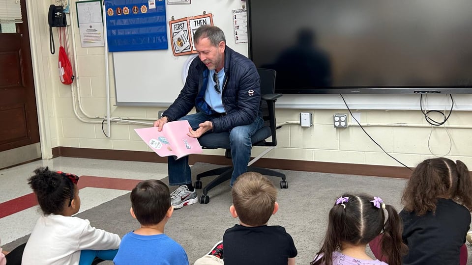 A man seated at the front of a classroom reads from pink papers to children sitting on the floor.