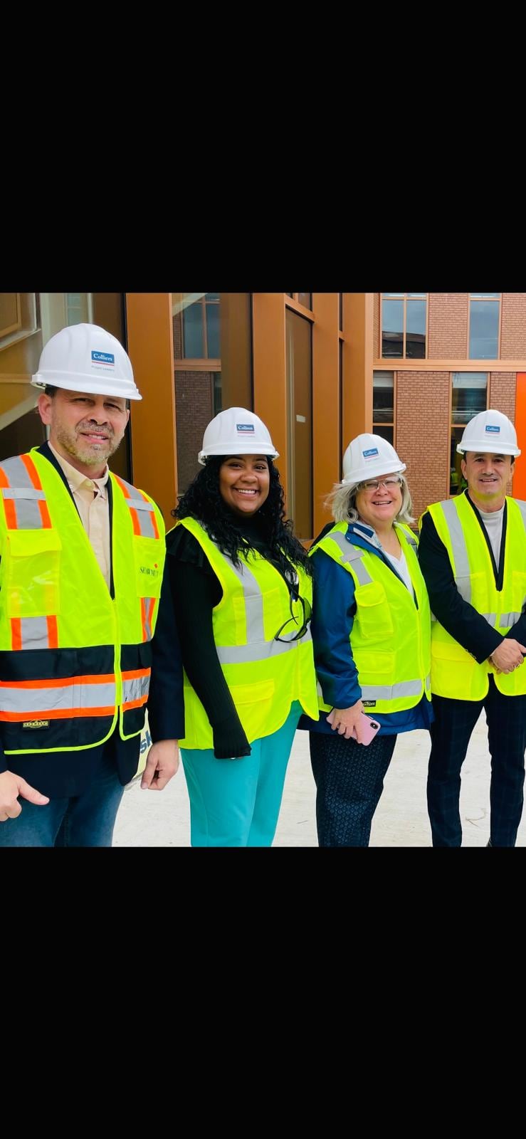 Four people wearing white hard hats and high-visibility vests pose together indoors.