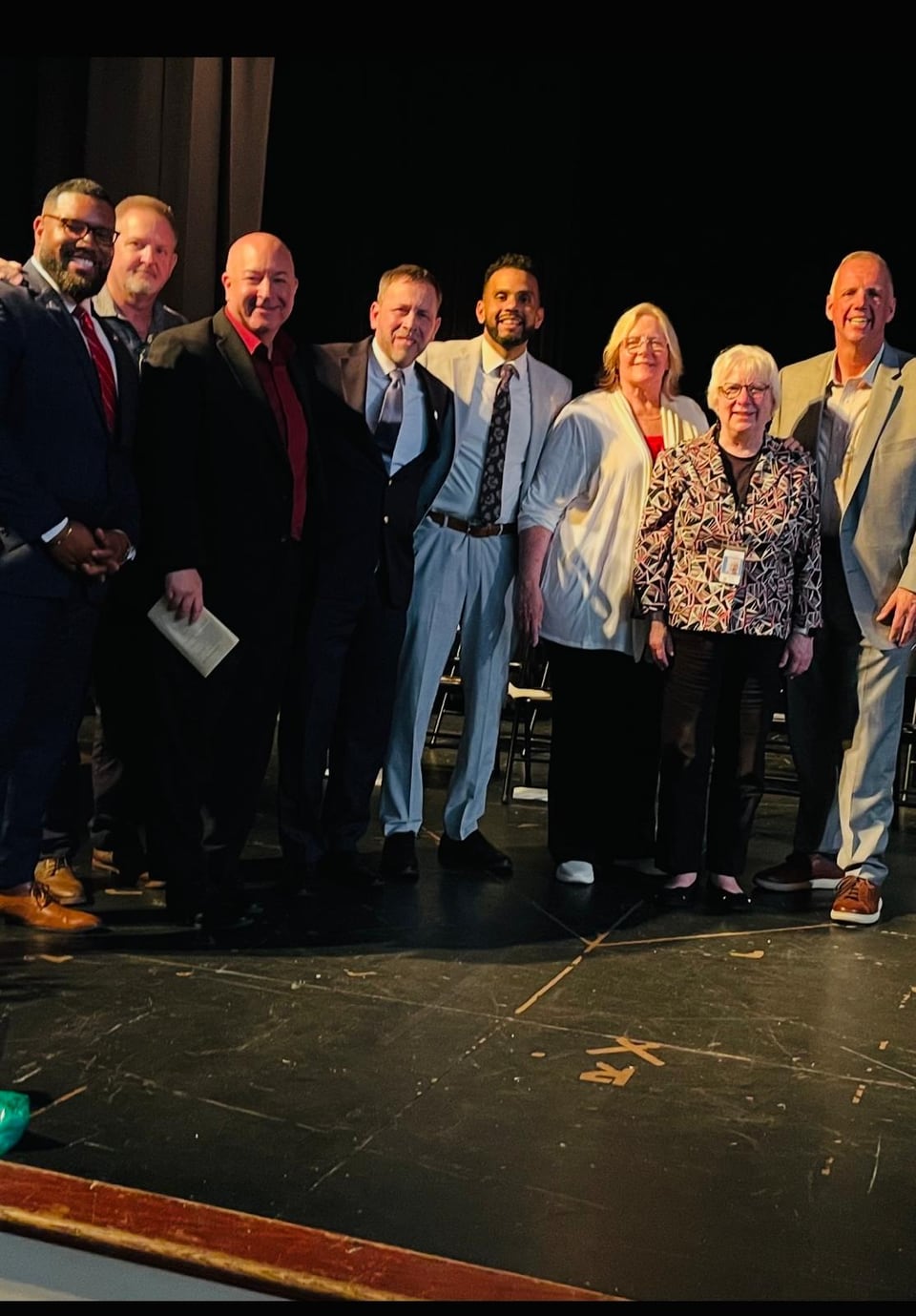A group of adults stands together and smiles on a dark stage.