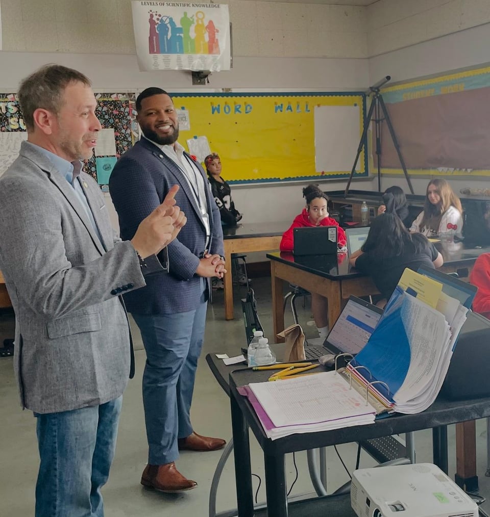 A man in a gray jacket speaks into a microphone in a classroom as another man and children look on.