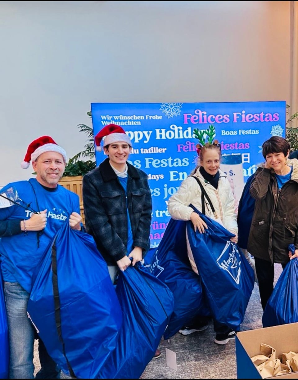 Four people in Santa hats and blue shirts pose with large blue bags in front of a holiday message.