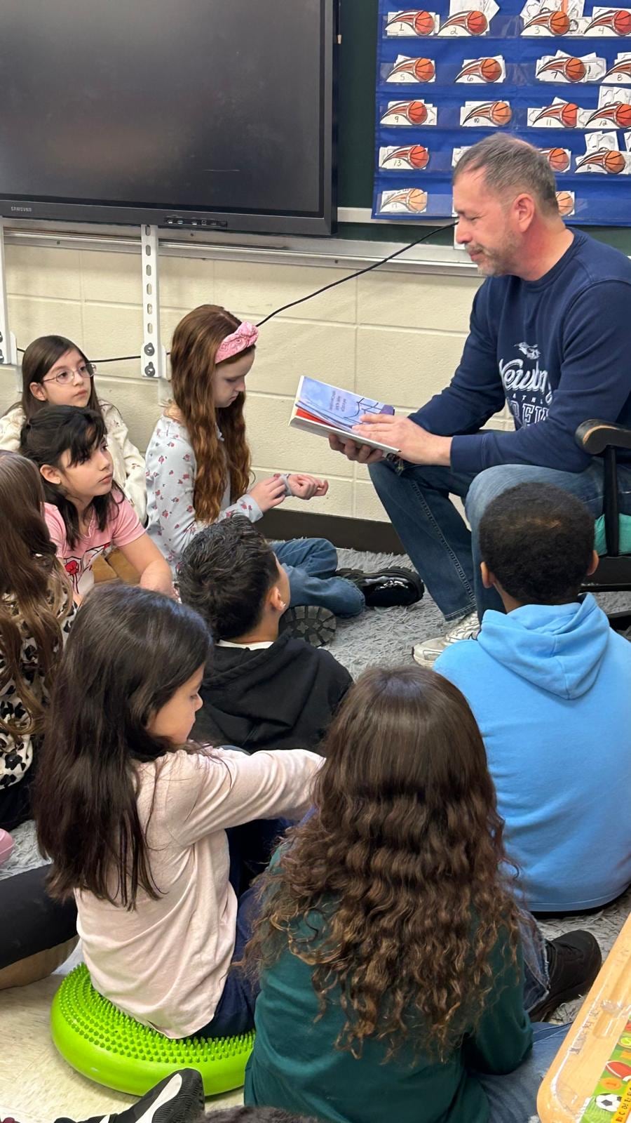 A seated man shows an open colorful book to children gathered on the classroom floor.