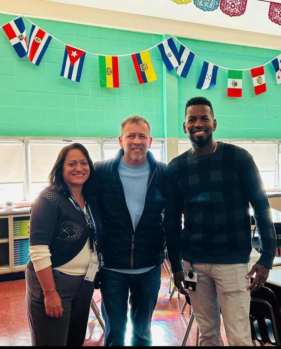 A woman and two men pose in a classroom beneath a row of international flags.