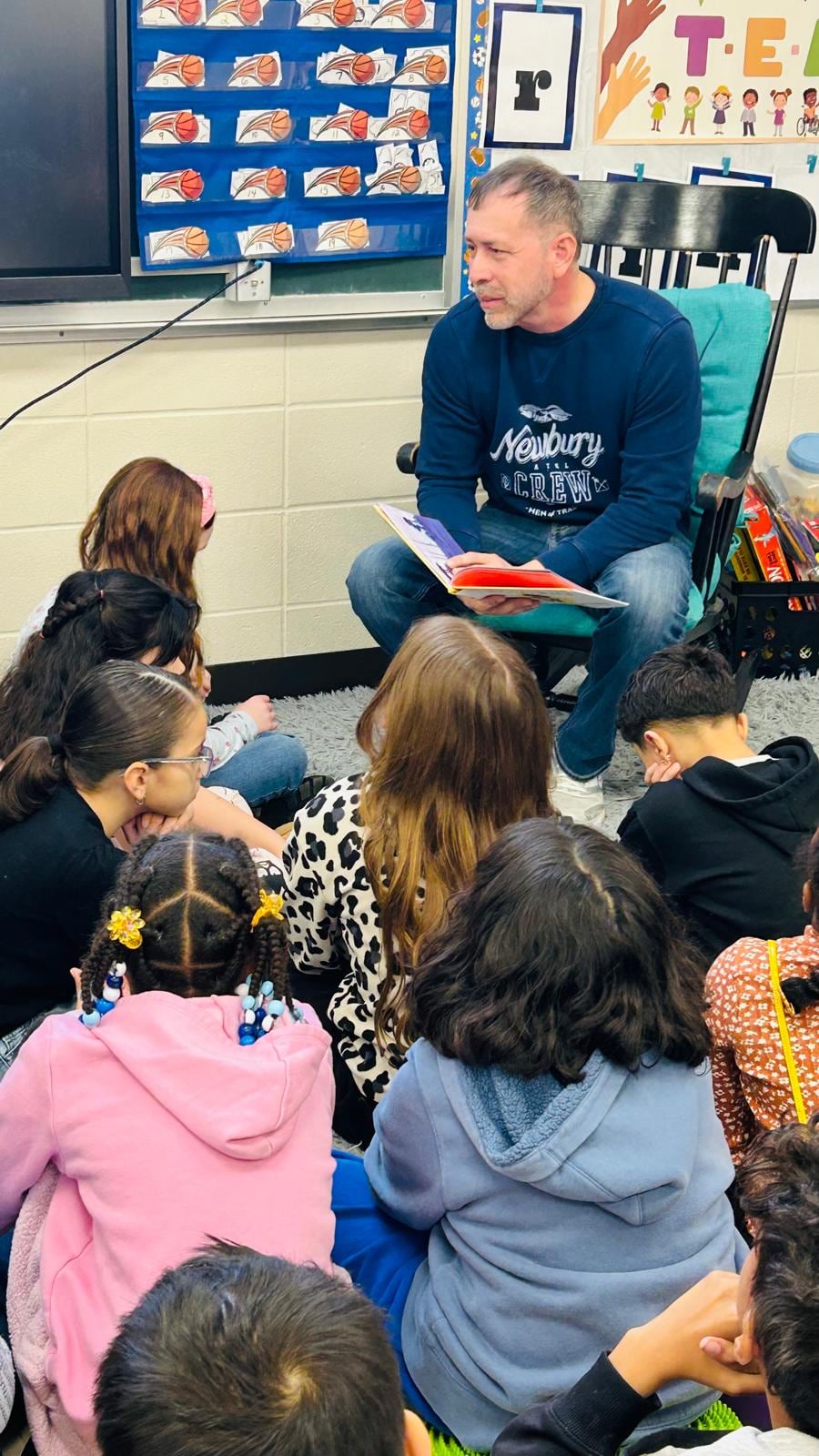 A man seated at the front of a classroom holds an open book while children sit around him on the floor.