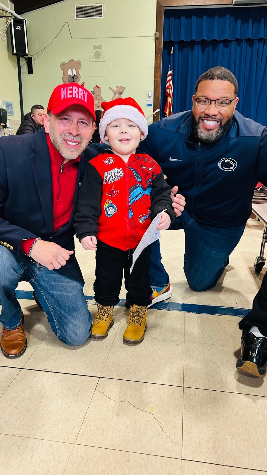 Juan Pablo Barrera and a bearded man kneel beside a child wearing a Santa hat and red Spider-Man vest.