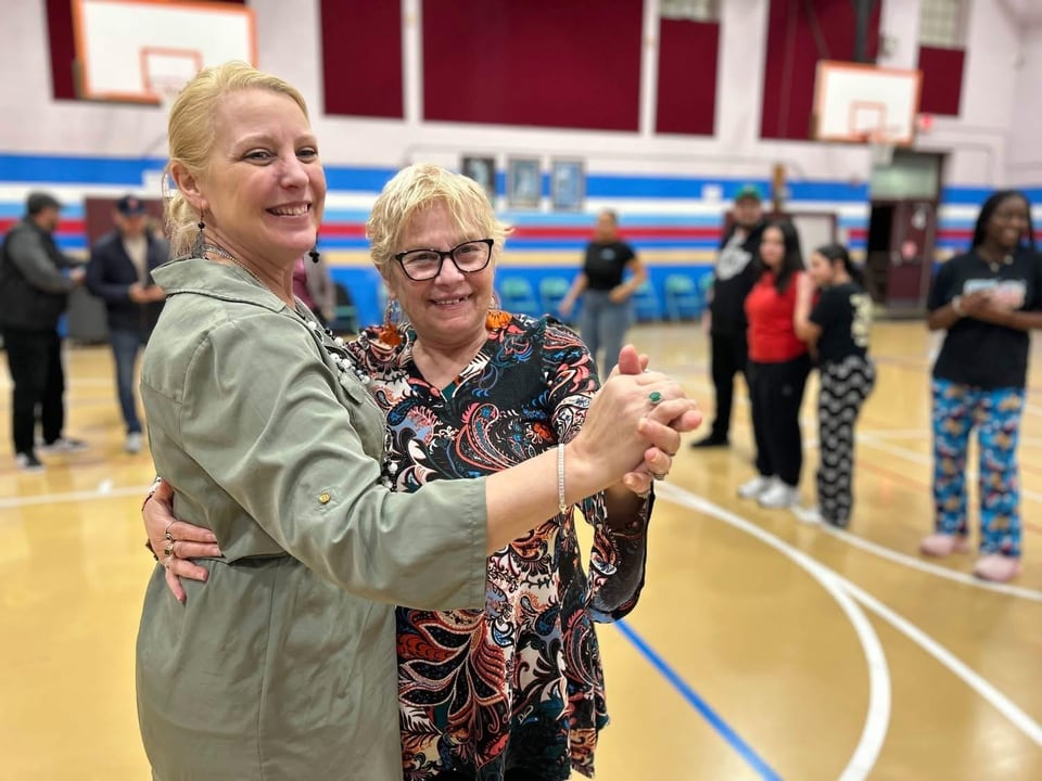 Two women hold hands while dancing in a gym as a group watches behind them.