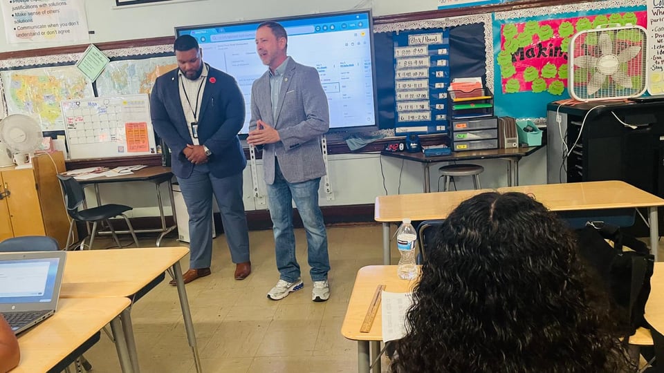 Juan Pablo Barrera gestures beside a suited man at the front of a classroom with desks and a display.