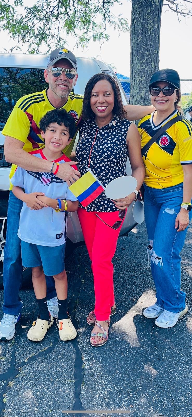 Four people pose outdoors by a parked vehicle; two adults wear yellow soccer jerseys, and one woman holds a small Colombian flag.