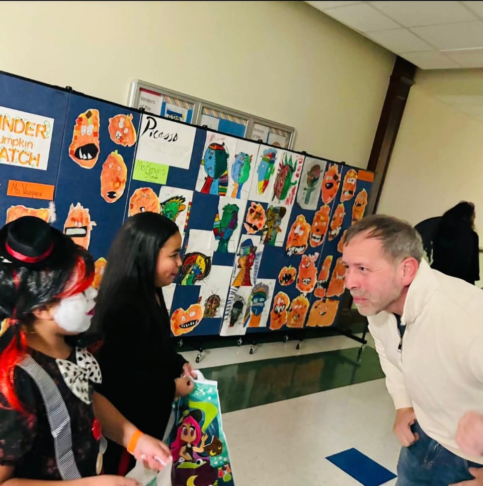 Juan Pablo Barrera leans toward two costumed children beside a colorful classroom display.