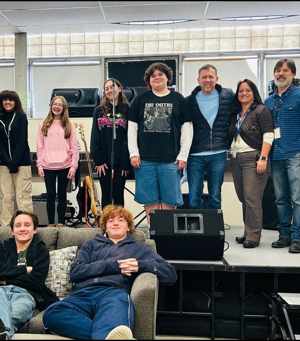A mixed-age group poses around microphones, guitars, speakers, and a couch in a music room.