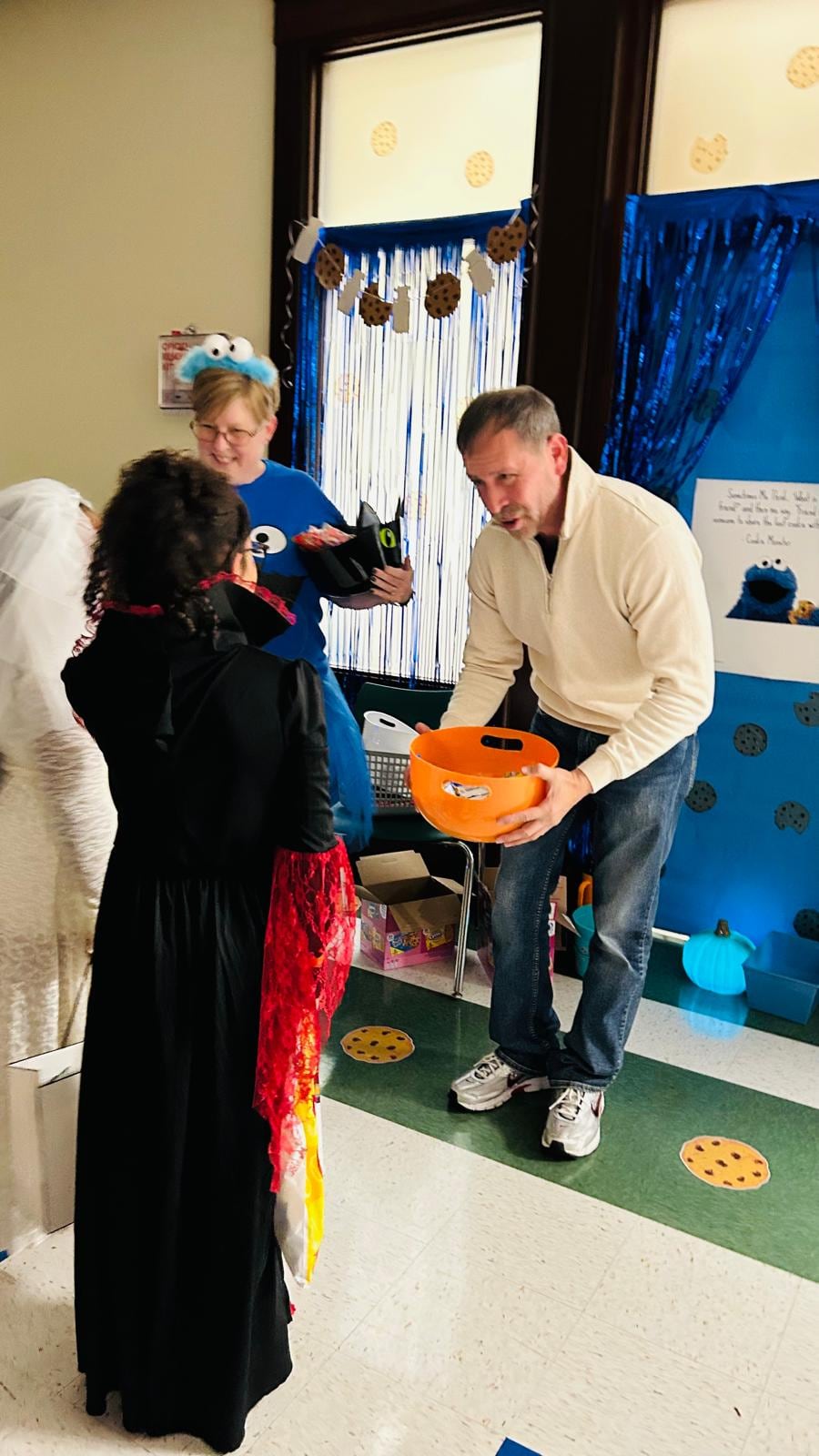 Juan Pablo Barrera holds an orange bucket toward a child in costume beside blue cookie-themed decorations.