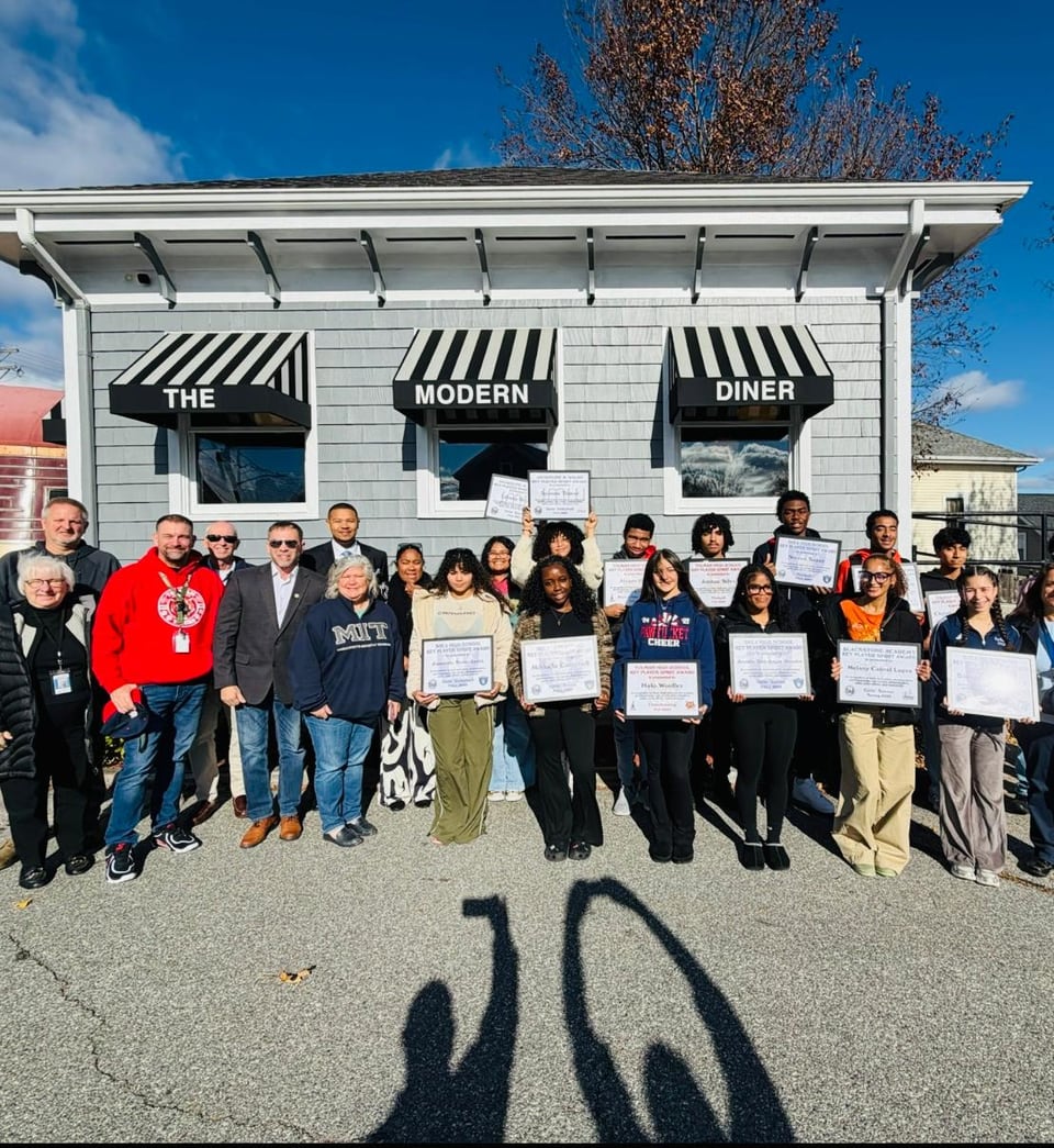 A large group poses outdoors before a diner, with several people holding framed certificates.
