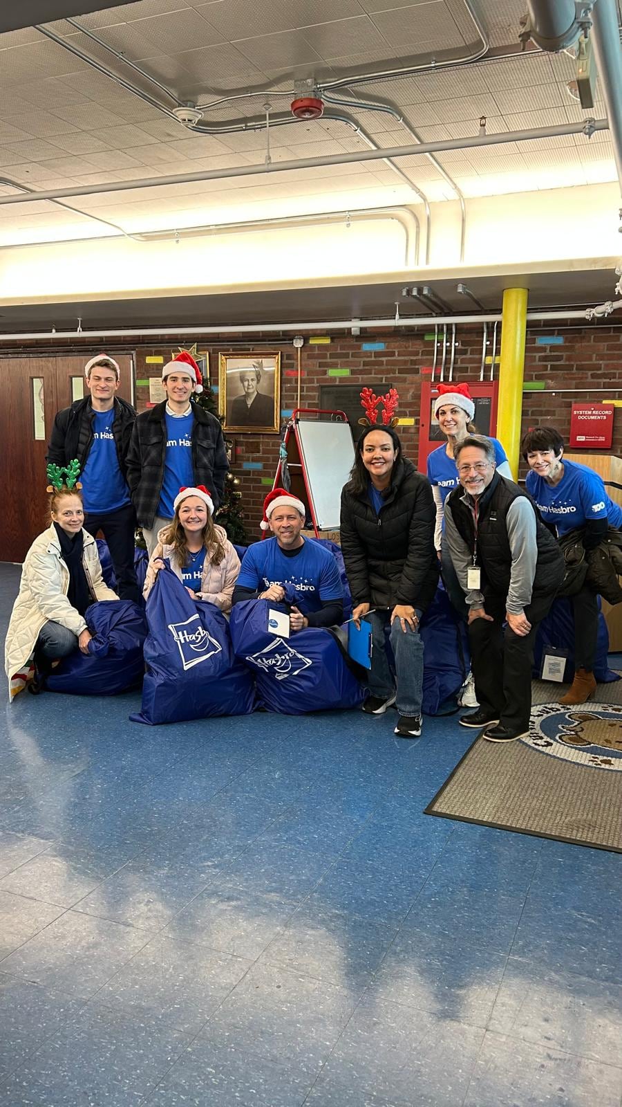 Ten adults in blue shirts, several wearing festive hats, pose indoors with large blue Hasbro bags.