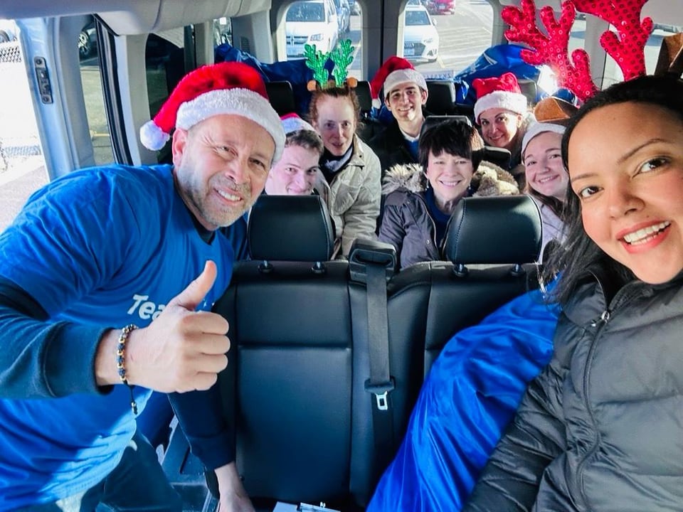 A man in a Santa hat gives a thumbs-up during a bus selfie with smiling passengers behind him.
