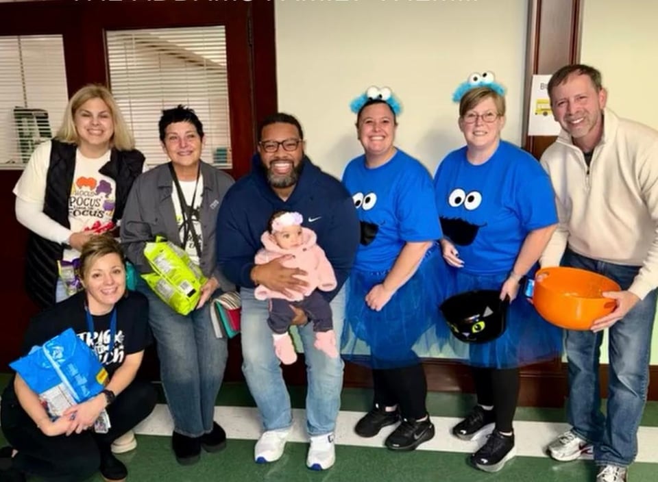 Seven adults and a baby pose indoors; two women wear blue character costumes and Juan Pablo Barrera holds an orange bucket.