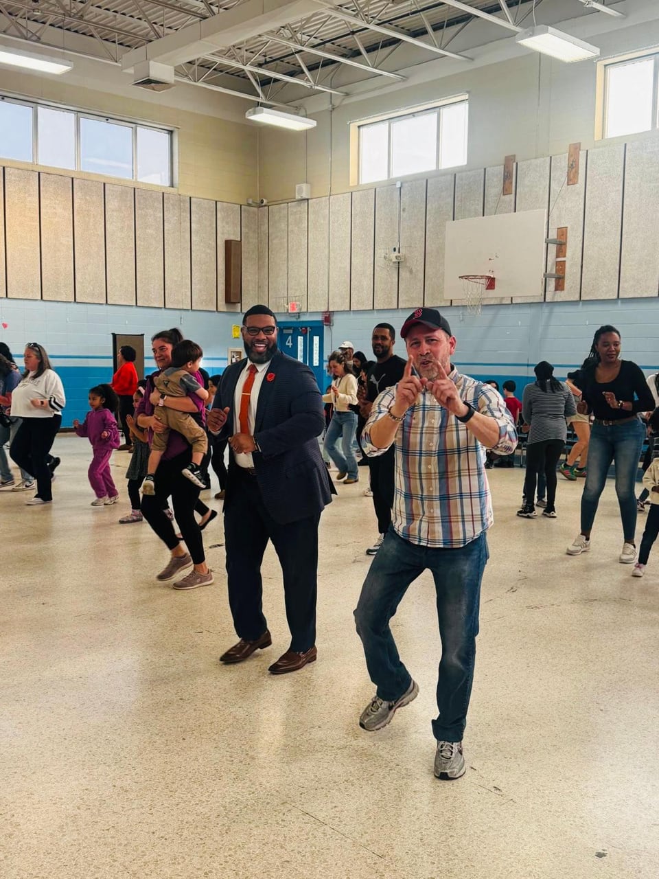 Juan Pablo Barrera points both index fingers while a suited man smiles and people dance behind them in a gym.