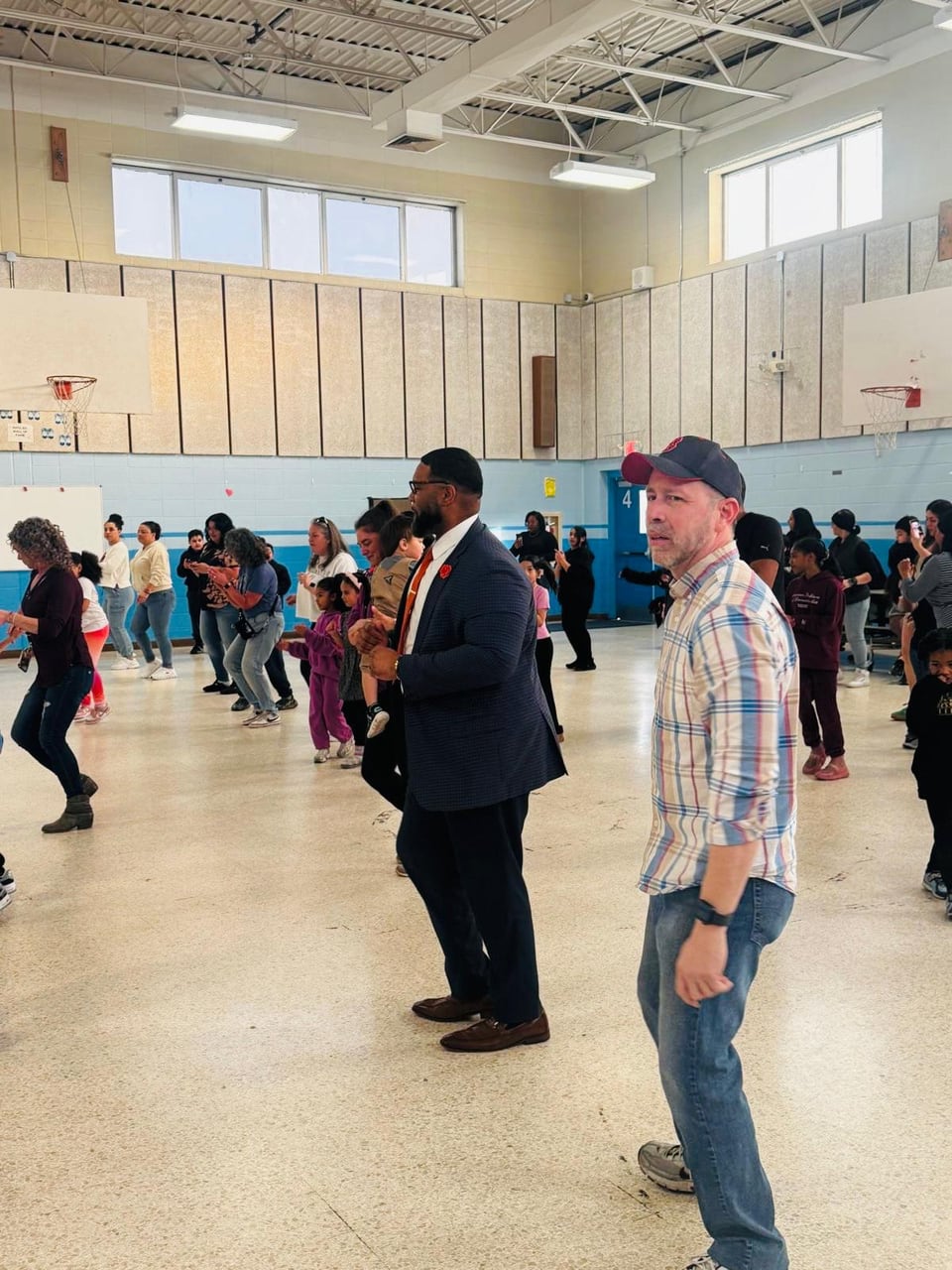 Juan Pablo Barrera and a suited man stand in profile among people dancing in a gym.
