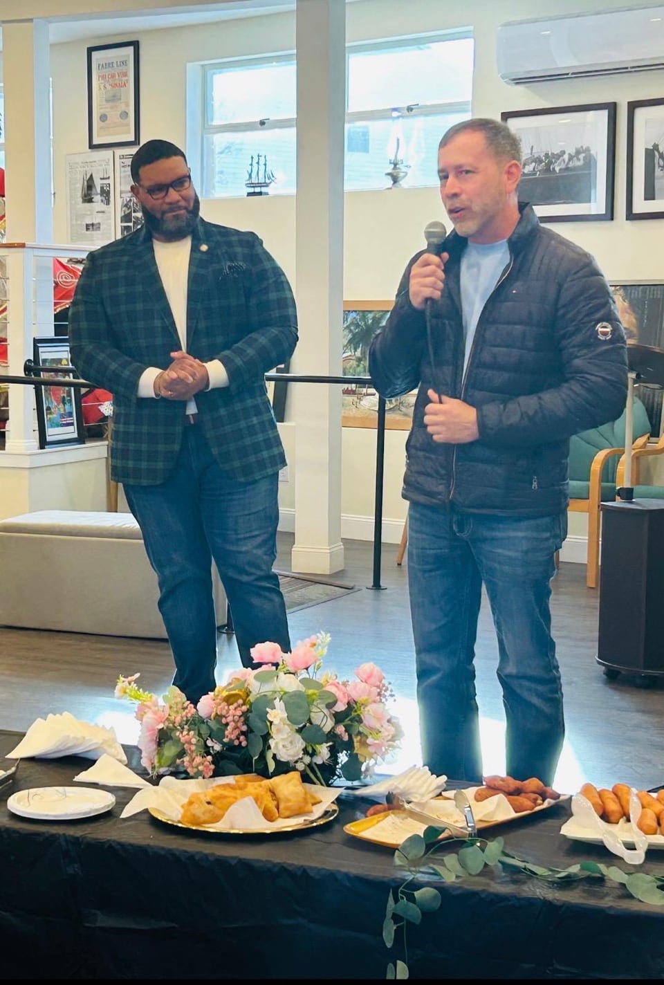 Juan Pablo Barrera speaks into a microphone beside a man, with flowers and trays of food on a table.