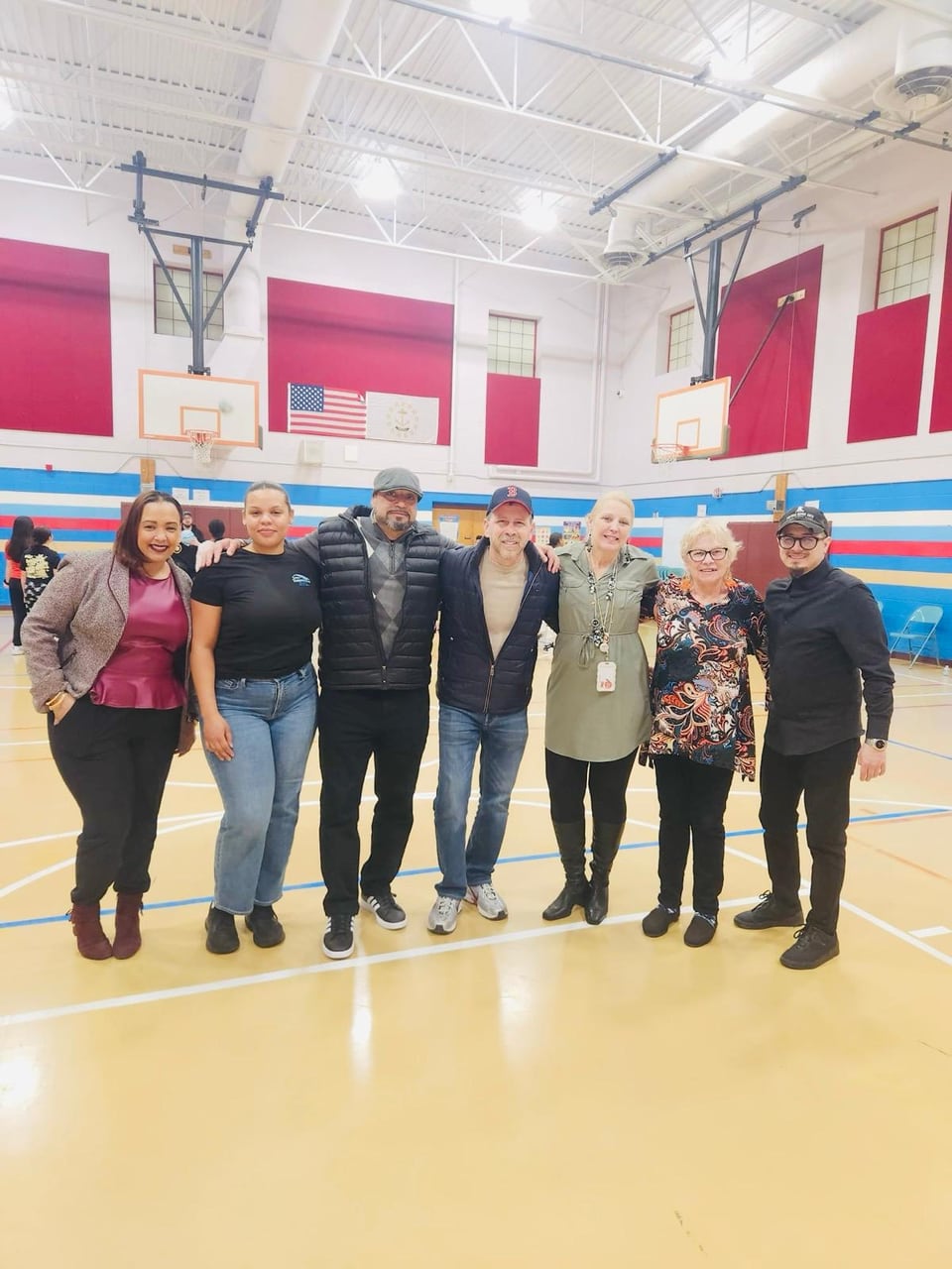 Seven adults stand arm in arm on a gym floor beneath basketball hoops and flags.
