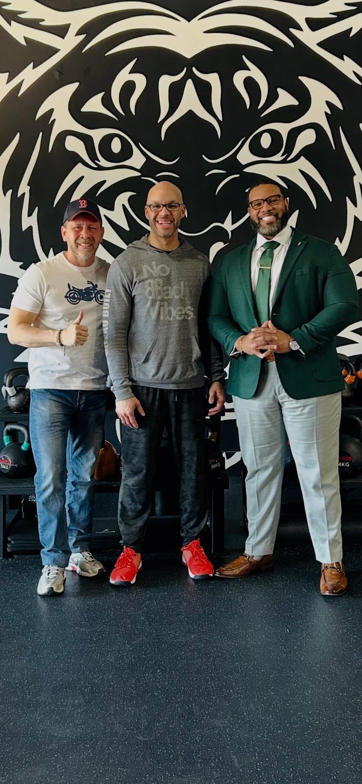 Juan Pablo Barrera poses with two men before a large black-and-white tiger mural and kettlebells.