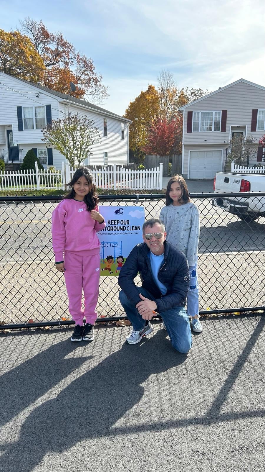 Juan Pablo Barrera crouches with two children by a fence and a 'Keep Our Playground Clean' sign.