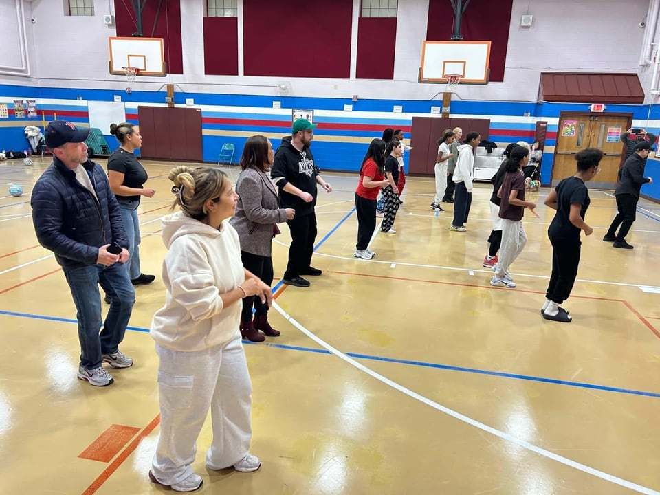 Adults stand in loose rows across a gym floor while a woman in white faces the group.