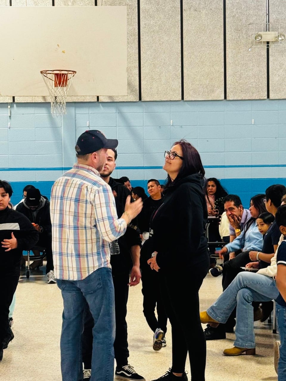 Juan Pablo Barrera in a plaid shirt and cap faces a woman with seated people behind them in a gym.