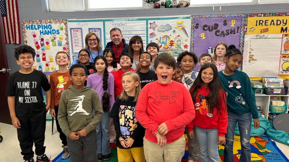 Children and three adults pose in a classroom before bulletin boards labeled Math, Reading, and Ready.