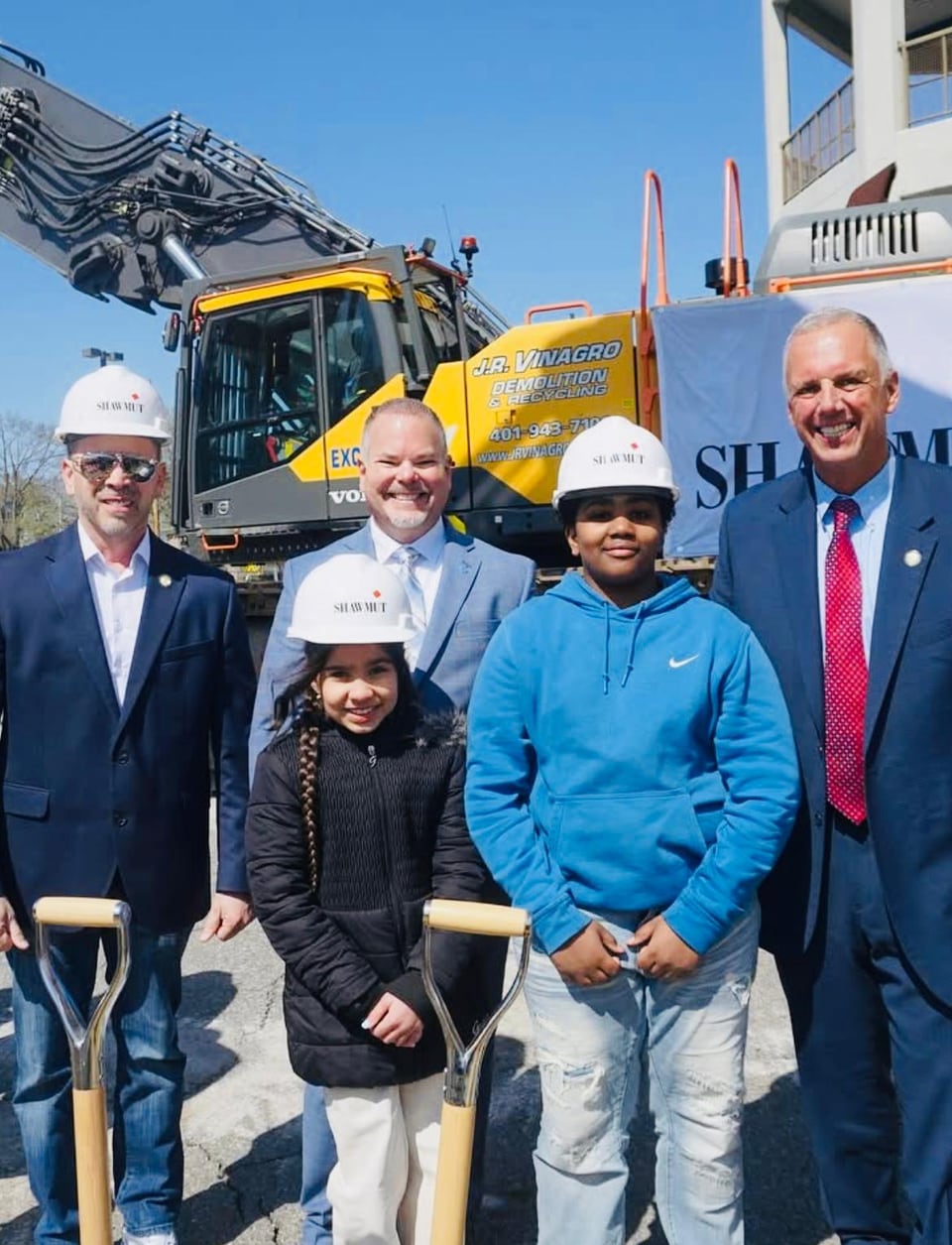 Juan Pablo Barrera stands with four people holding shovels in front of a yellow excavator.