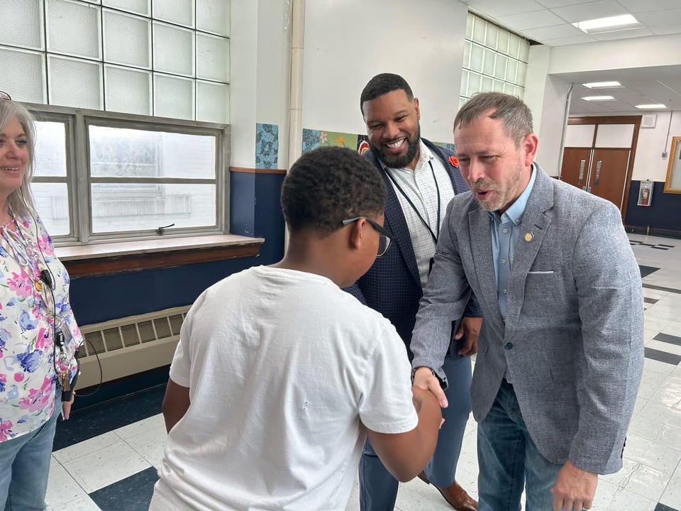 Juan Pablo Barrera leans forward to shake hands with a young person in a hallway as others look on.