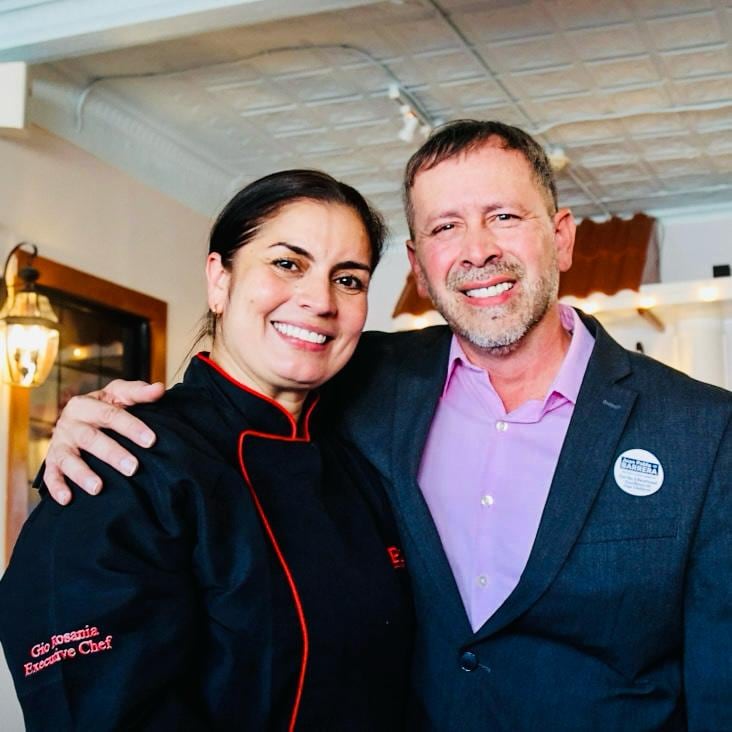 Juan Pablo Barrera poses with a smiling woman in a black jacket with red piping.