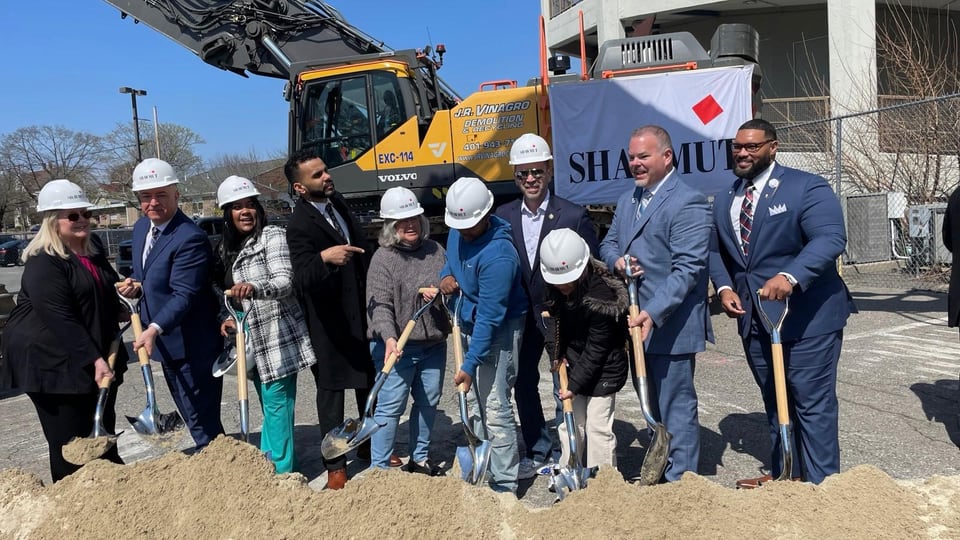 A large group wearing hard hats holds shovels in a dirt lot beside a yellow excavator.