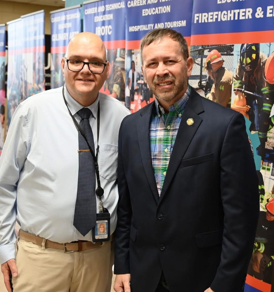 Juan Pablo Barrera poses with a man in a white shirt and tie in front of a colorful printed banner.
