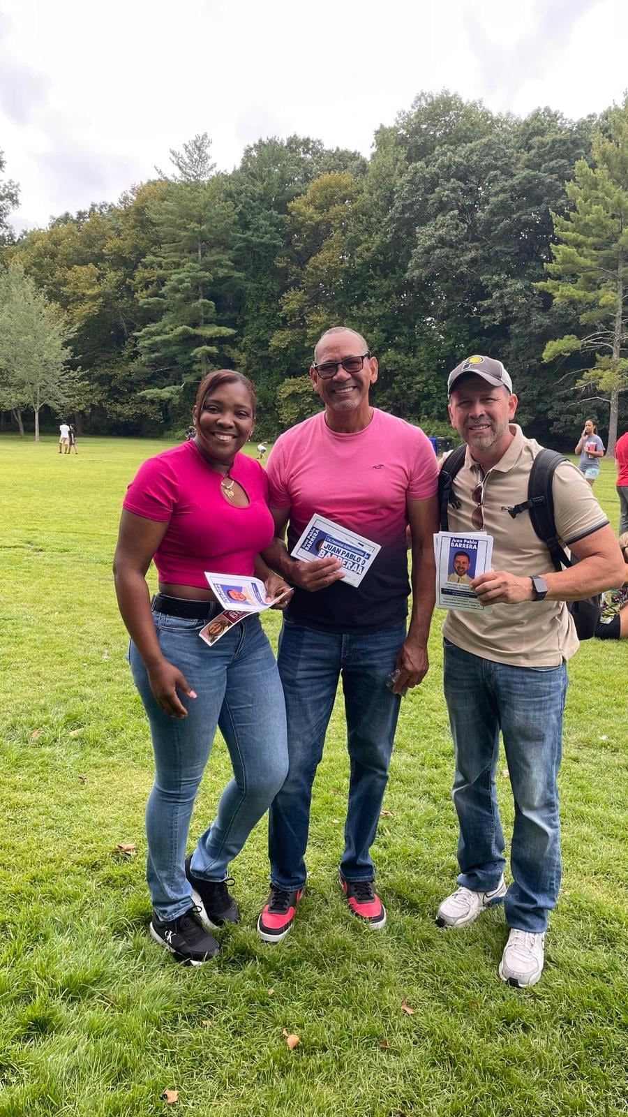Three adults stand together on a grassy field with trees in the background.