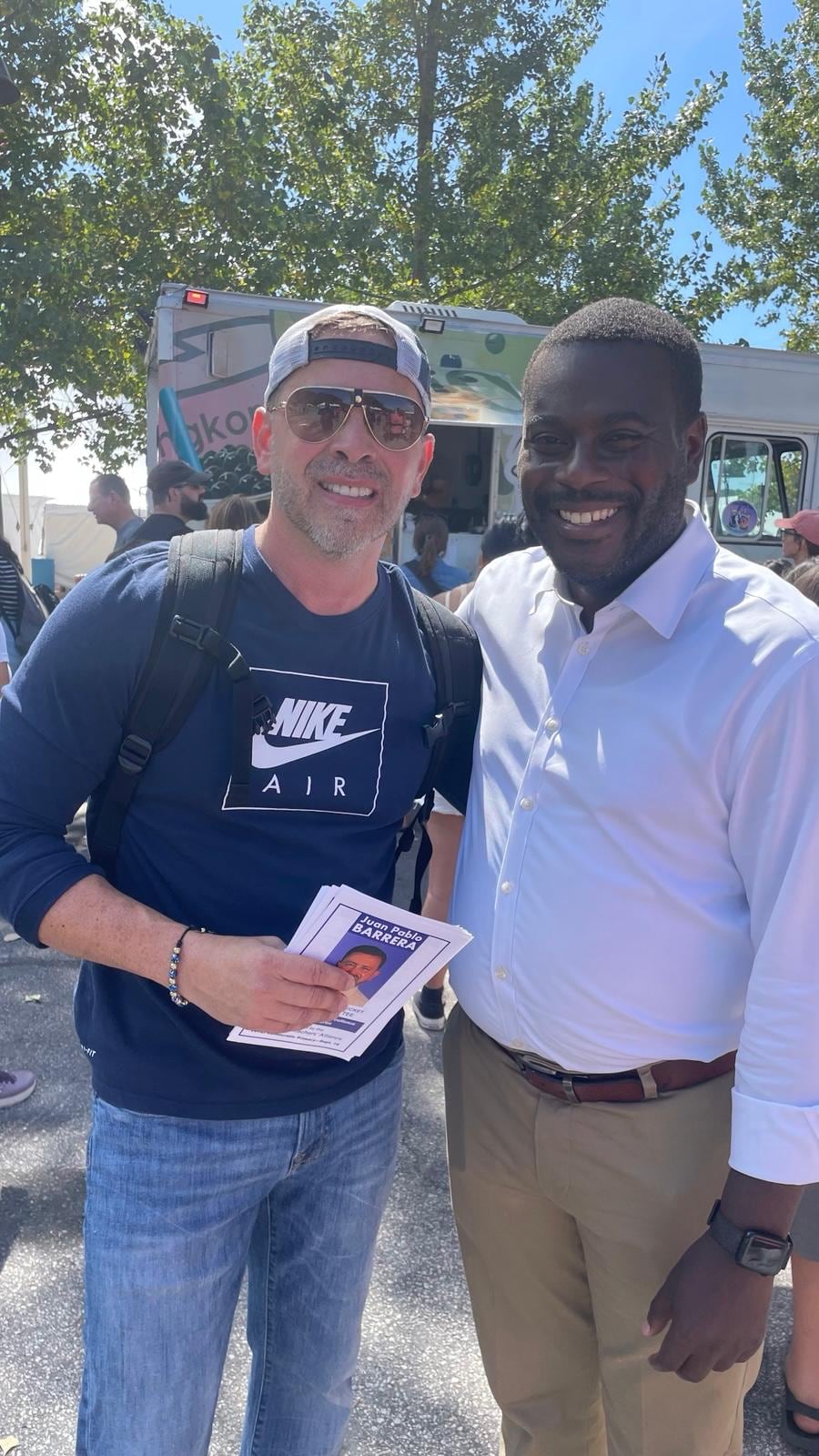 Juan Pablo Barrera stands shoulder to shoulder with a smiling man outdoors while holding printed material.